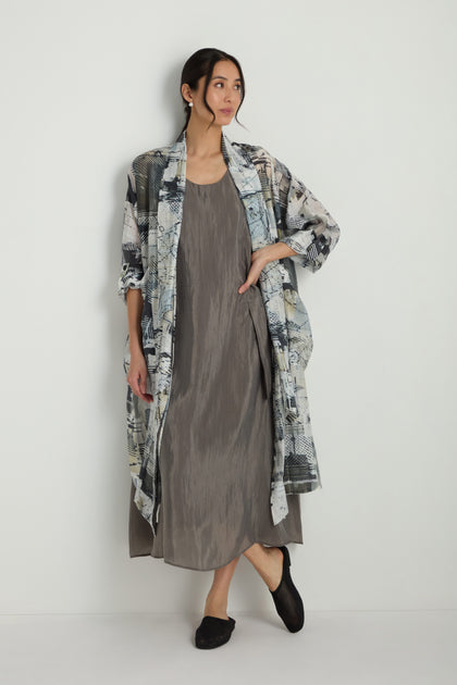 A woman stands against a plain wall wearing a loose, sleeveless gray dress, the Shaded Print Long Coat, and black shoes.