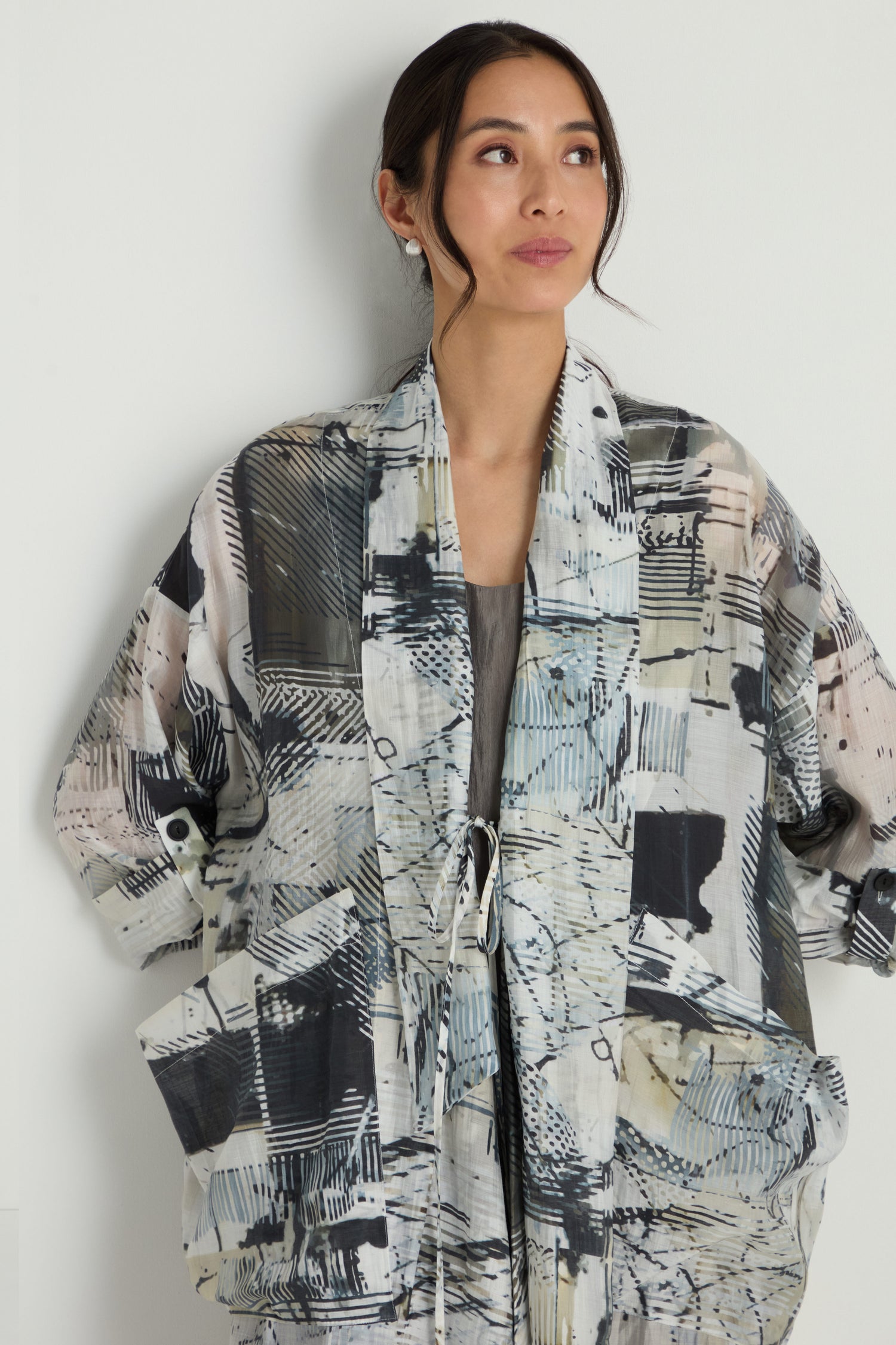 A woman stands against a white wall wearing the Shaded Print Long Coat, featuring a loose fit, large pockets, and an abstract shaded print in black, white, and gray.