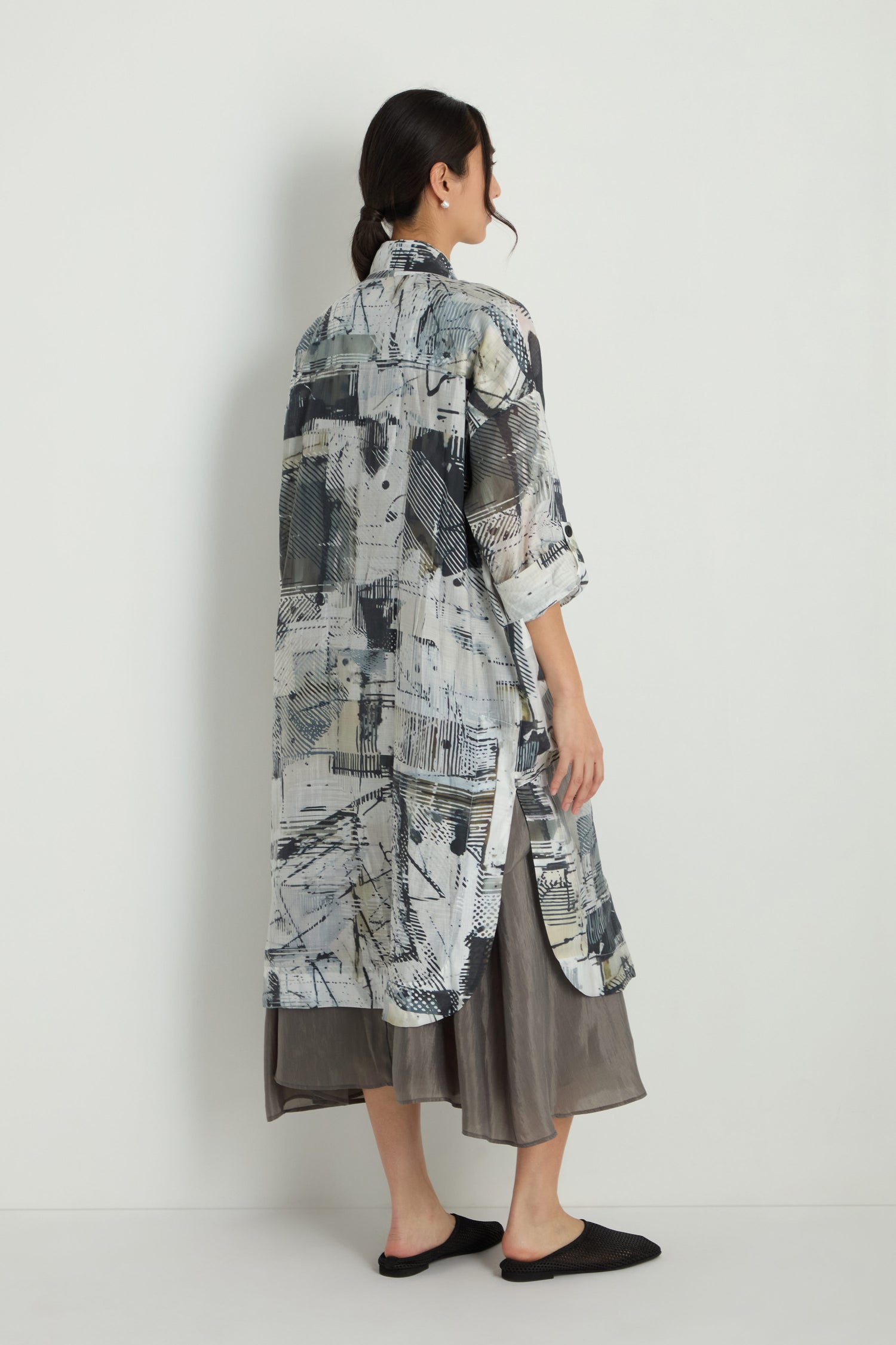 A woman stands sideways facing a wall, wearing the Shaded Print Long Coat over a gray dress and black slip-on shoes.