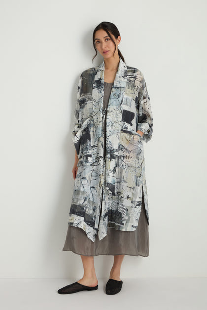 A woman stands against a white wall wearing the Shaded Print Long Coat, a kimono-style jacket in a soft tencel blend, layered over a loose gray dress with black slip-on shoes.