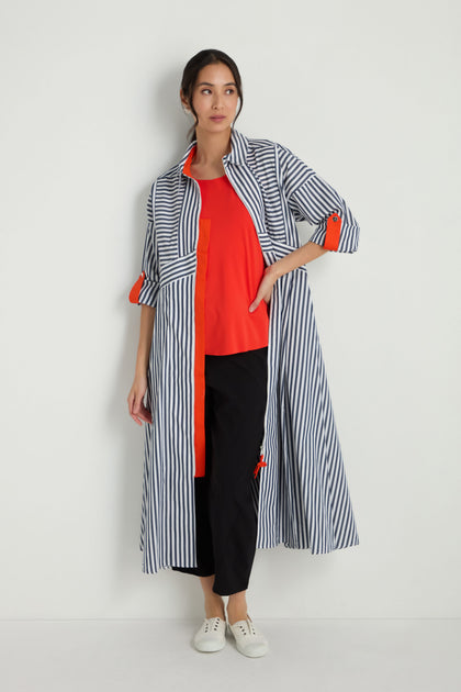 A woman stands against a white wall wearing the Striped Zip Dress in blue and white, layered over a red top with black pants and white sneakers.