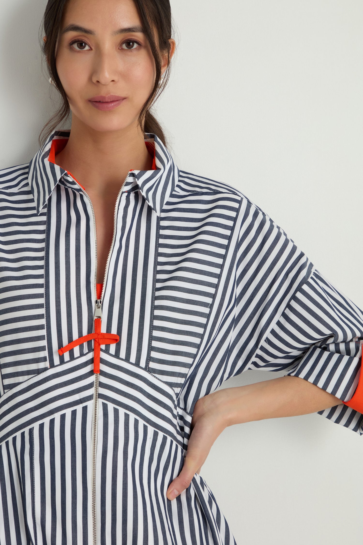 A woman wears the Striped Zip Dress, a cotton midi with navy and white vertical and diagonal stripes, orange accents, a collared neckline, fit-and-flare silhouette, and front zip closure, standing against a plain background.