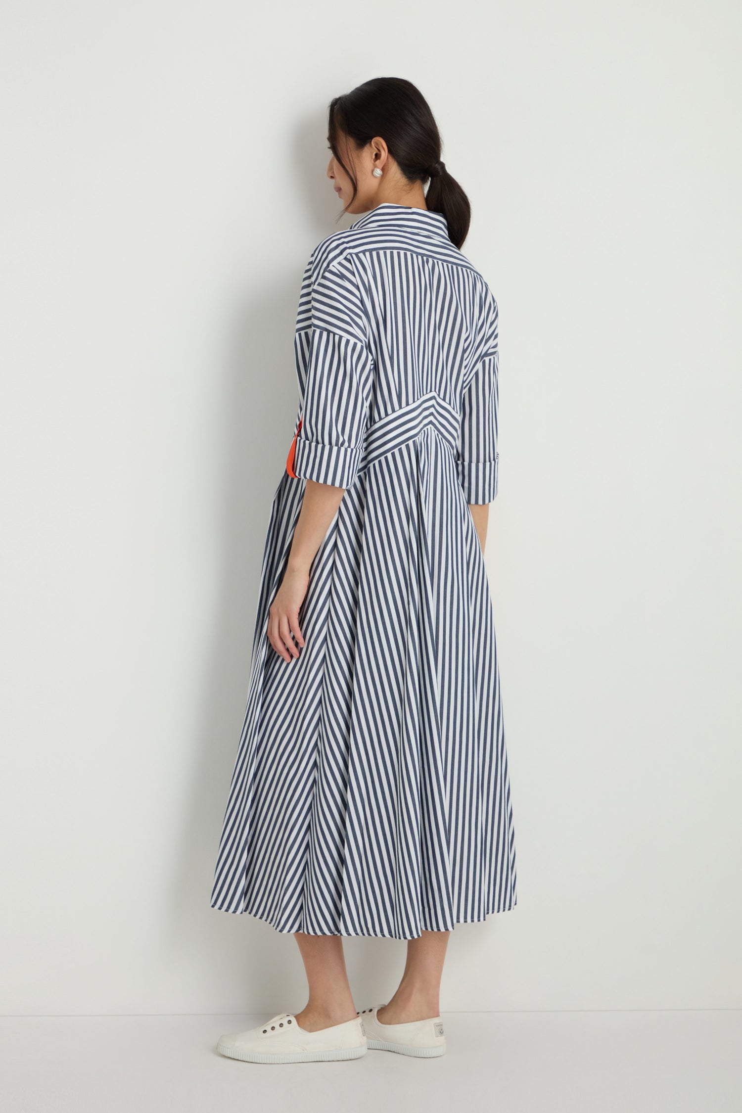 A woman faces away, wearing the Striped Zip Dress—a blue and white vertical striped cotton midi with three-quarter sleeves—paired with white sneakers, set against a plain white background.