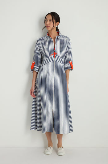 Model wears the Striped Zip Dress featuring blue and white stripes, red accents, and a flattering fit and flare silhouette, paired with white sneakers against a plain white background.