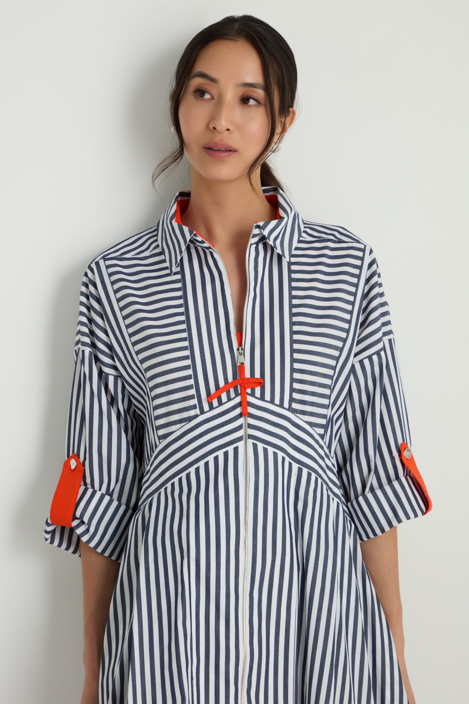 A woman wears the Striped Zip Dress, a blue and white cotton midi featuring orange accents on the collar and sleeves, standing against a plain white background.
