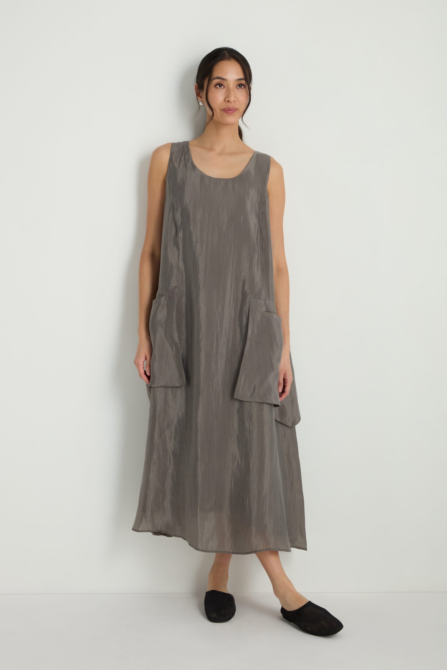A woman stands against a white wall wearing the Sleeveless Pocket Dress in gray, featuring an A-line silhouette and large front pockets, paired with black slip-on shoes.