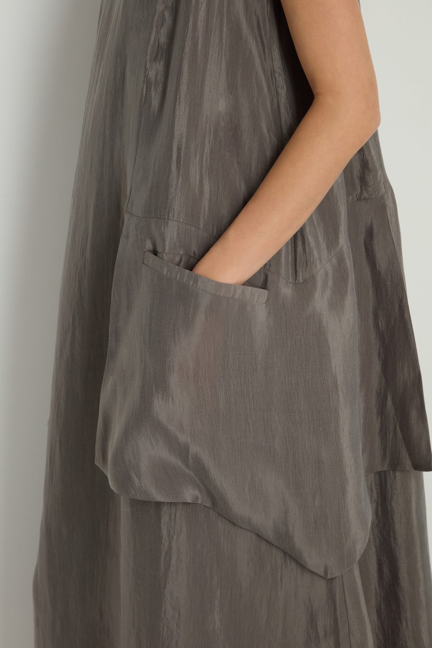 Model stands against a light wall wearing a loose, shiny gray Sleeveless Pocket Dress, arm bent with hand tucked into the large side pocket.