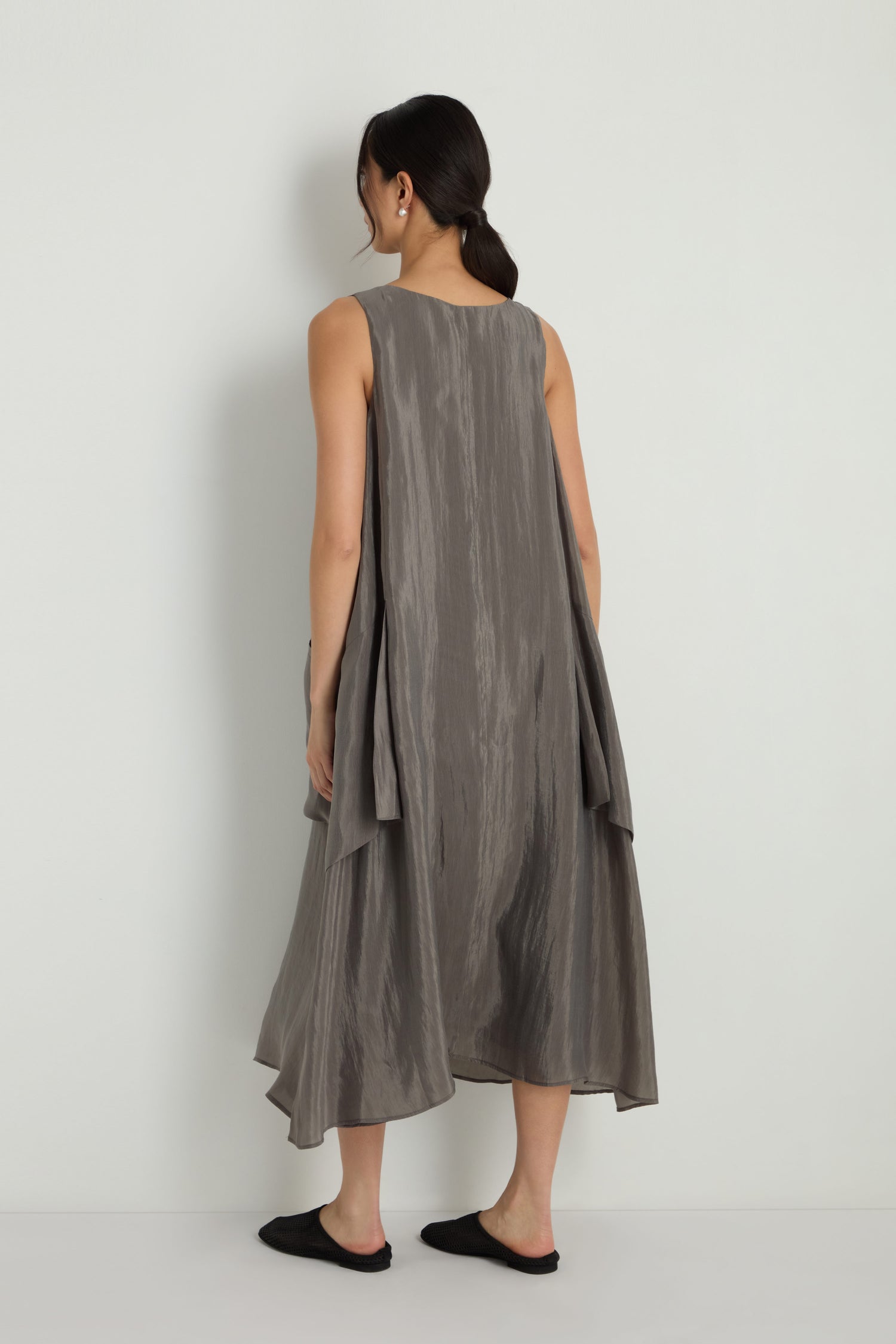 A woman stands facing away in the Sleeveless Pocket Dress, a loose-fitting gray midi with pockets and textured finish, styled with black flats against a plain white background.