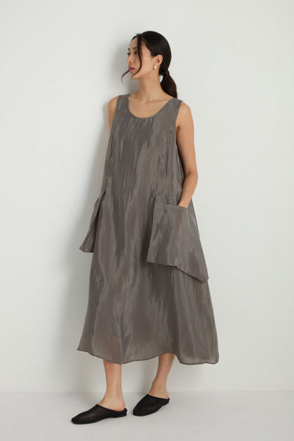 A woman stands against a plain white wall wearing the Sleeveless Pocket Dress featuring an A-line silhouette and oversized pockets, paired with black slip-on shoes and hoop earrings.