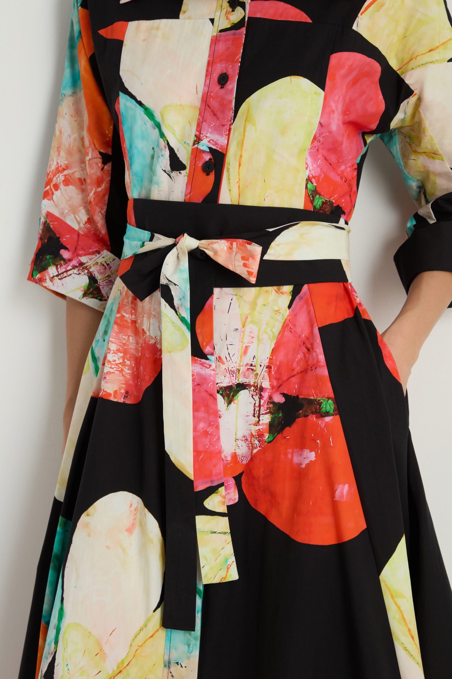 Model wears the Bright Floral Flared Dress, a black cotton midi with vibrant floral prints, buttoned top, matching fabric waist tie, and pockets—one hand casually placed in the pocket.