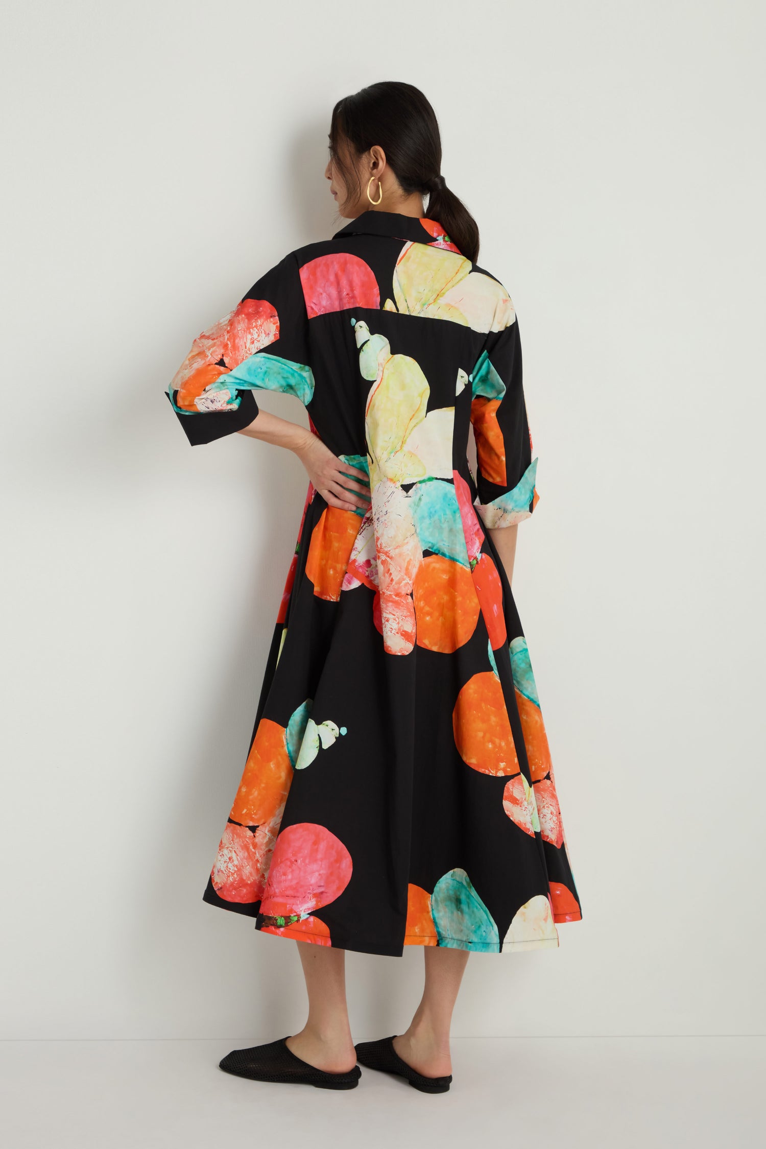 A woman faces away, wearing the Bright Floral Flared Dress—a black cotton midi dress with orange, teal, and yellow abstract circular patterns—standing against a plain white background.