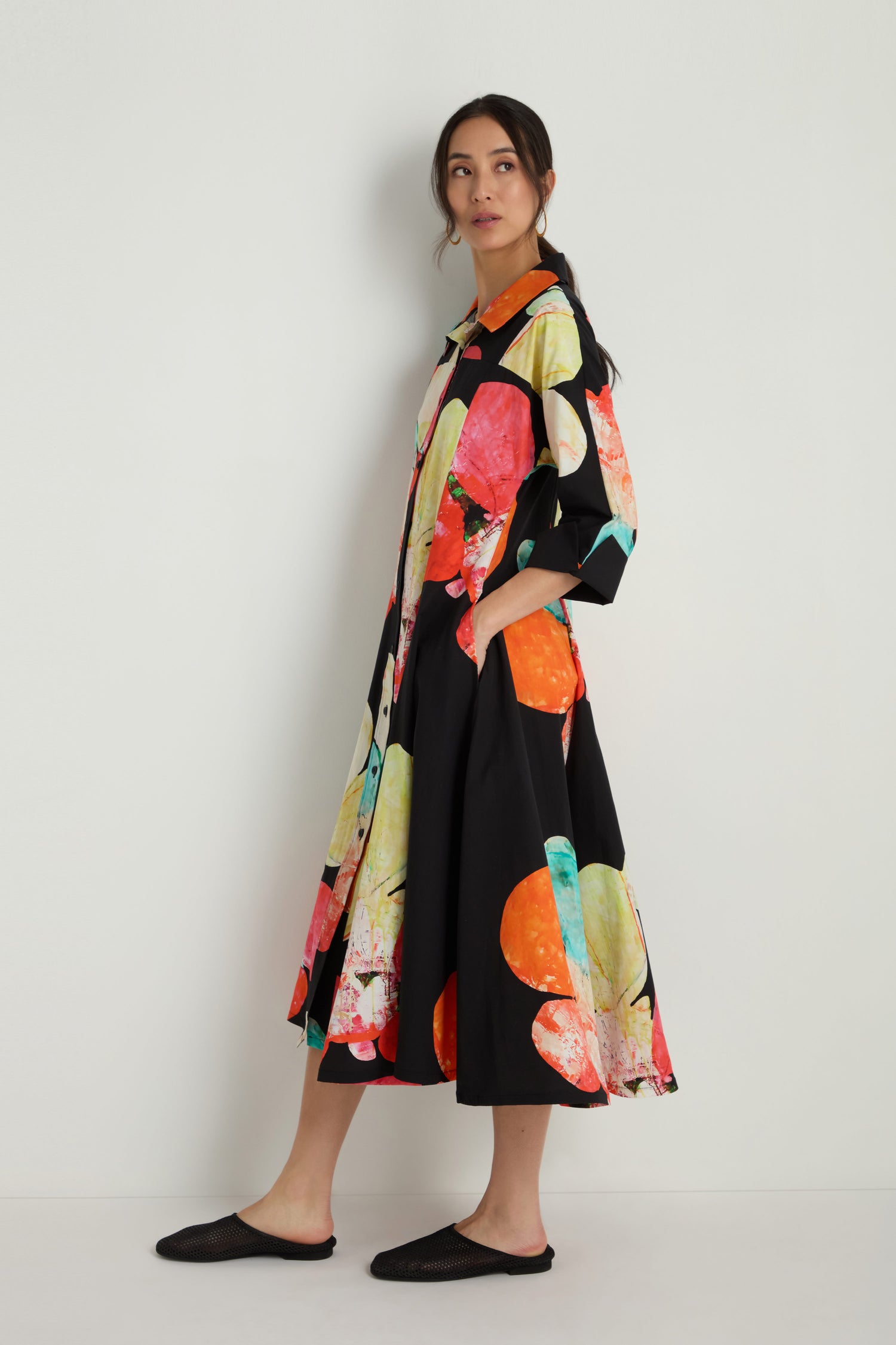 A woman stands sideways against a white wall, wearing the Bright Floral Flared Dress featuring large, colorful abstract patterns and black flat shoes.