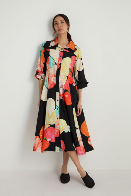 A woman stands against a white wall wearing the Bright Floral Flared Dress with a colorful abstract print and black slip-on shoes.