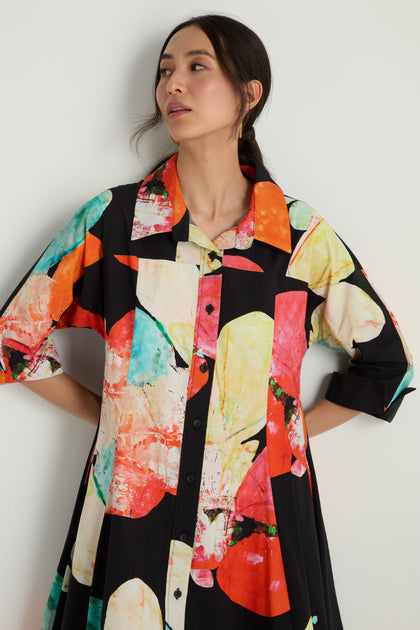 Woman wearing the Bright Floral Flared Dress, a loose cotton midi with bold red, orange, yellow, and teal abstract splashes on a black background.