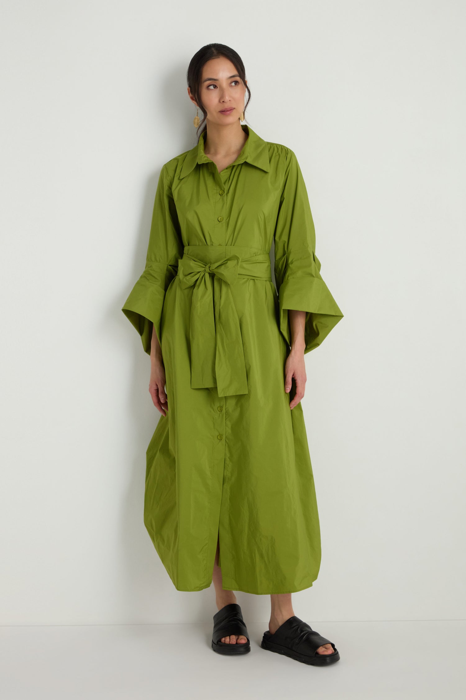 A woman wearing the Taffeta Button-Through Lime Dress and black sandals stands against a plain white wall.