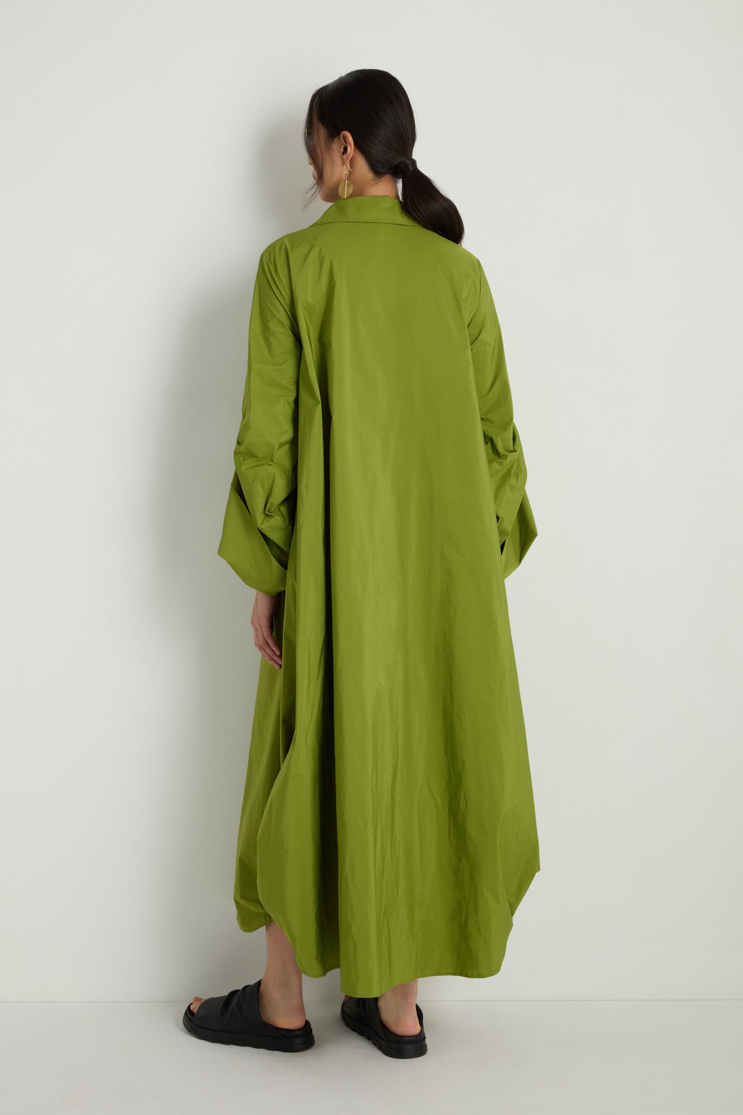 A person faces a white wall, wearing the Taffeta Button-Through Lime Dress styled as a coat, with black sandals, black pants, and their hair tied back.