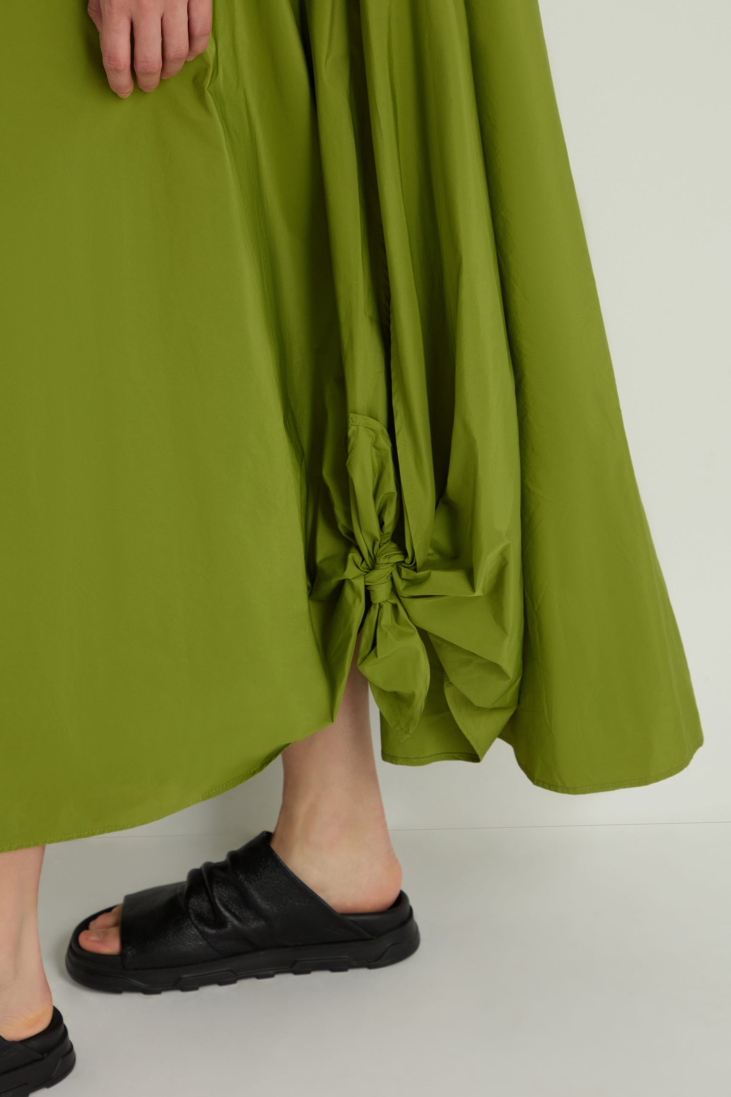 Close-up of a person wearing the Taffeta Button-Through Lime Dress with a gathered knot detail near the hem, paired with black slide sandals, standing on a white floor.