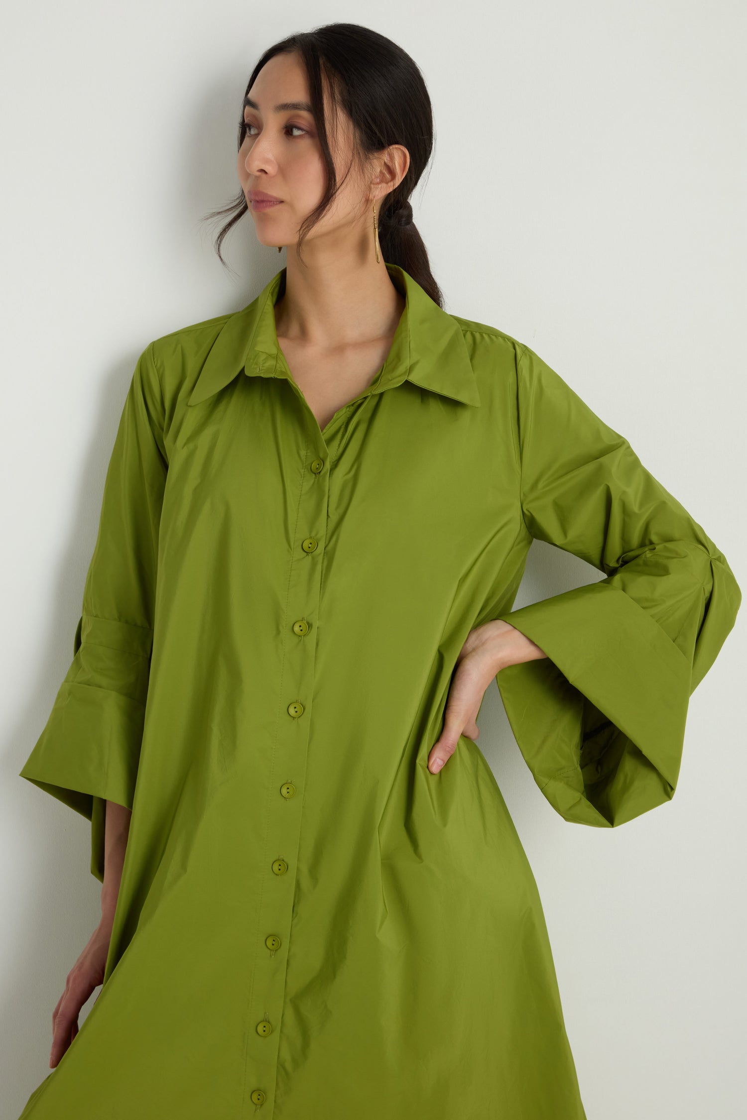 A woman wearing the Taffeta Button-Through Lime Dress, a loose-fitting long-sleeve outfit, stands against a plain white background with one hand on her hip.