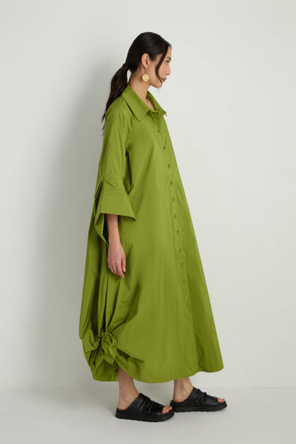 A woman stands sideways against a plain background wearing the Taffeta Button-Through Lime Dress with wide sleeves, a gathered hem detail, black sandals, and gold earrings.