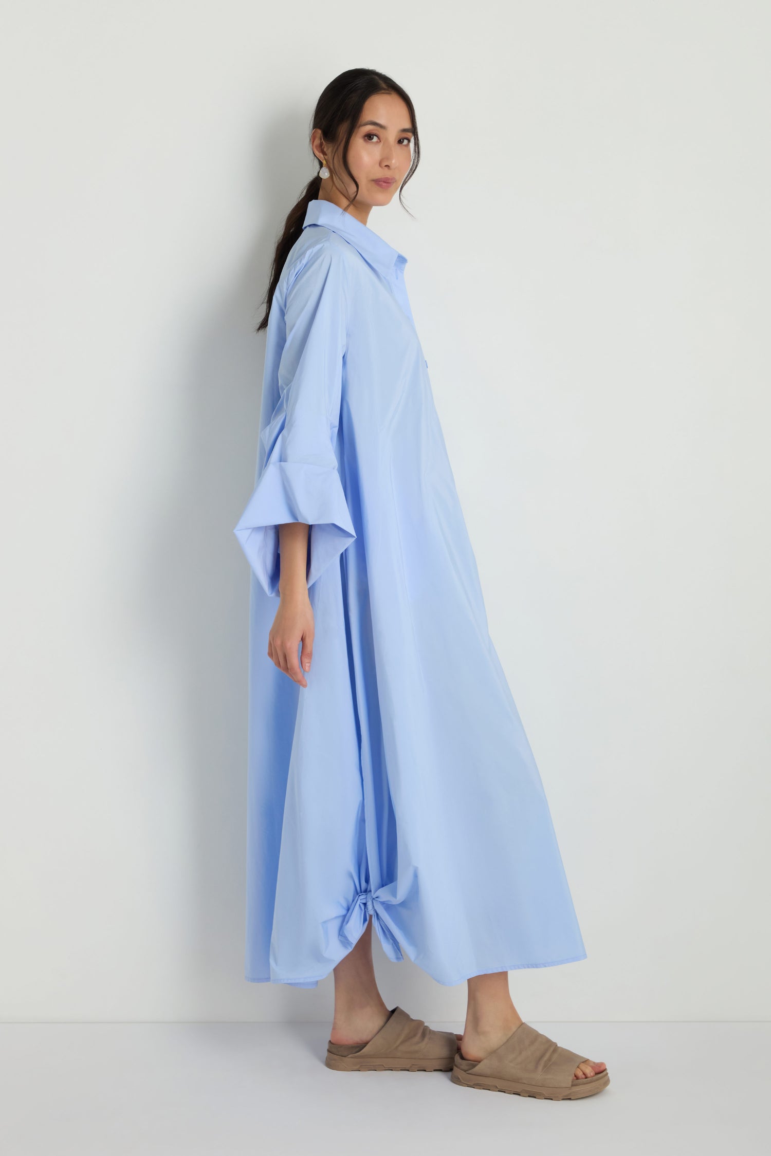A woman wears the Taffeta Button-Through Sky Dress, a light blue, midi-length, loose-fit style with wide sleeves, paired with tan slide sandals, standing sideways against a plain white background.