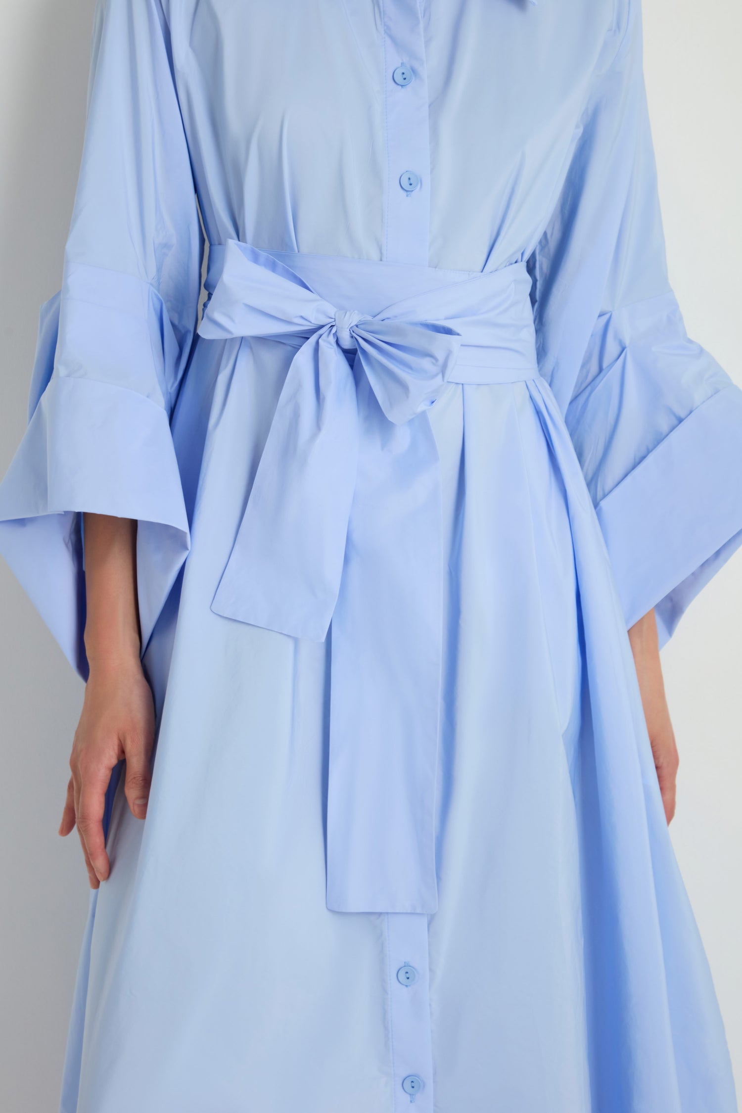 Person wearing the Taffeta Button-Through Sky Dress, a light blue, long-sleeved midi dress with wide sleeves and a matching fabric belt tied at the waist.