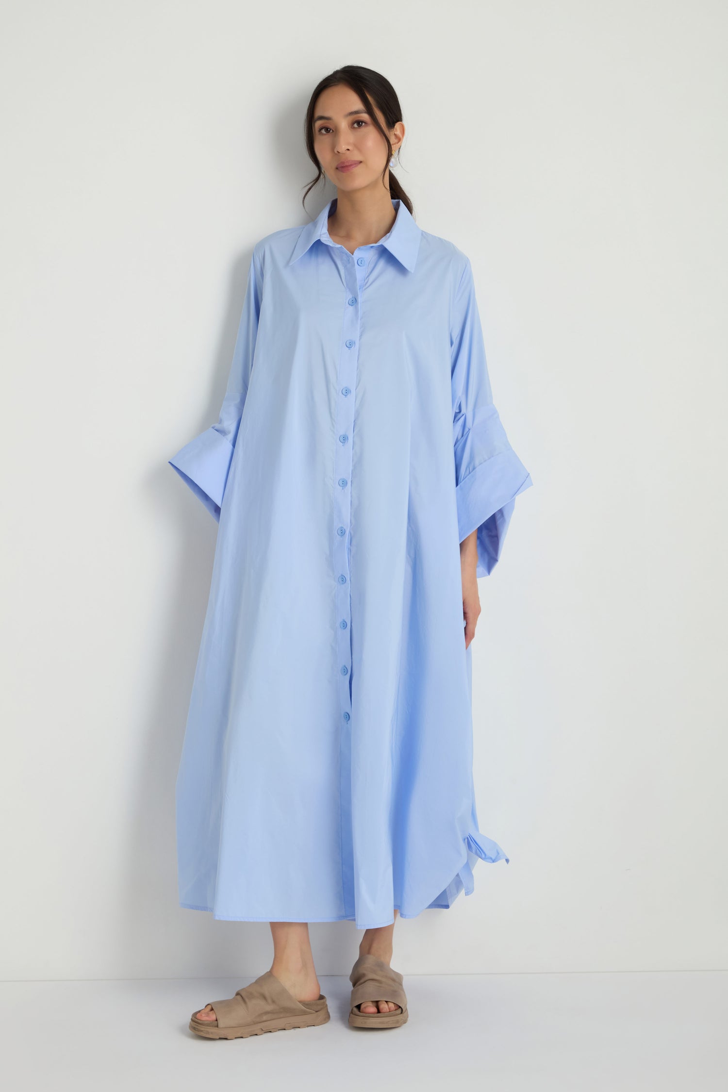 A woman stands against a white wall wearing the Taffeta Button-Through Sky Dress, a loose-fitting, midi-length light blue dress with wide sleeves, paired with beige slide sandals.