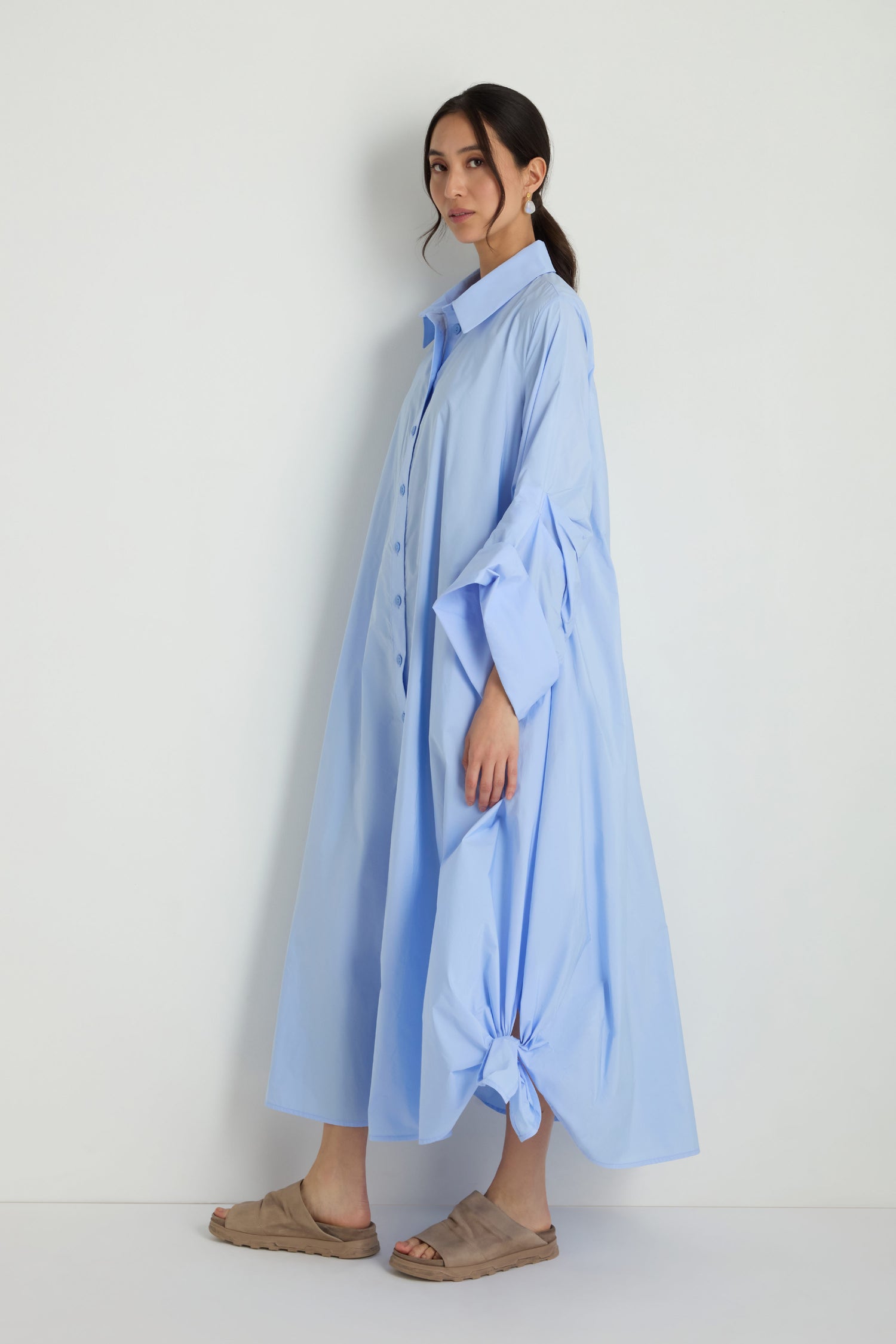 A woman stands against a plain background wearing the Taffeta Button-Through Sky Dress, a loose, light blue midi dress with side knots, paired with beige slip-on sandals.