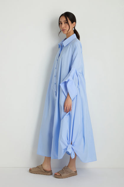 A woman stands against a plain background wearing the Taffeta Button-Through Sky Dress, a loose, light blue midi dress with side knots, paired with beige slip-on sandals.