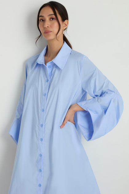 A woman stands against a plain white background wearing the Taffeta Button-Through Sky Dress, a loose-fitting, light blue midi dress with wide sleeves.