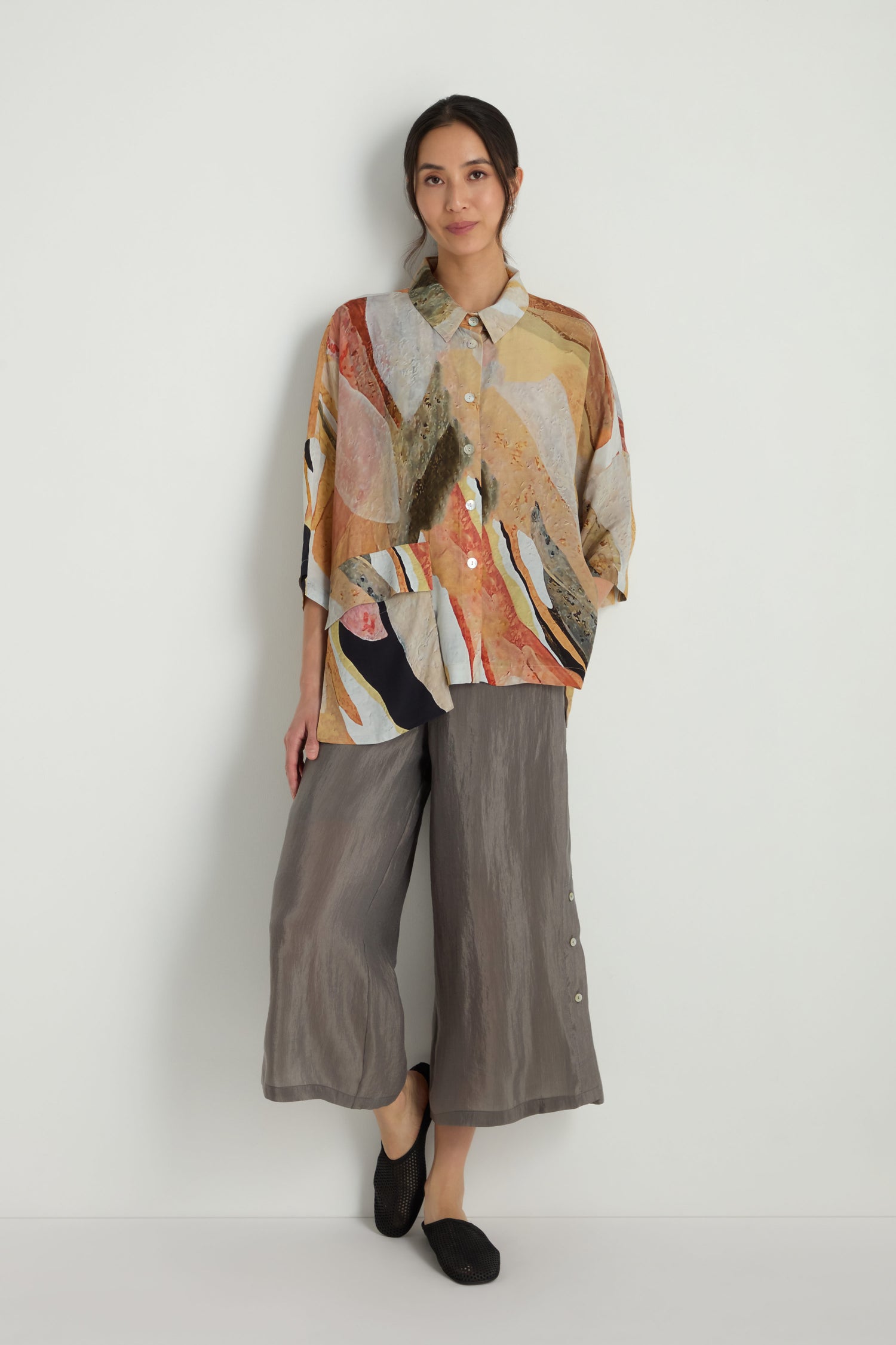 A woman stands against a plain wall, wearing the Abstract Earth Print Shirt in a relaxed silhouette with a soft viscose blend, paired with loose gray cropped pants and black shoes.