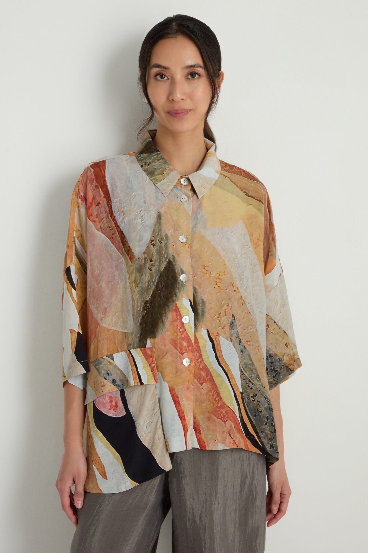A woman stands against a plain wall, wearing the Abstract Earth Print Shirt—a relaxed-fit top crafted from a soft viscose blend—paired with gray pants.