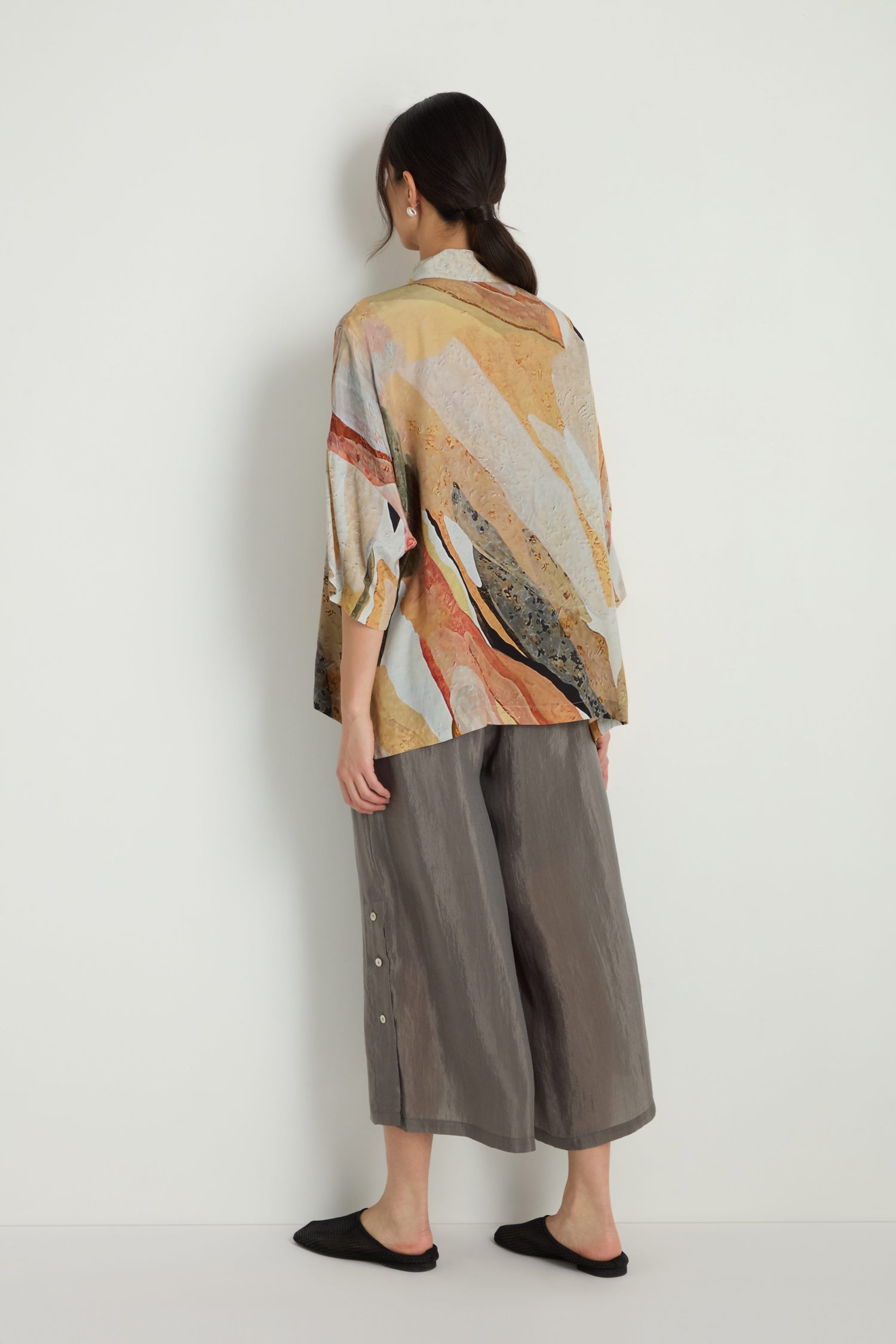 A woman faces a white wall wearing the Abstract Earth Print Shirt, featuring an abstract earth pattern in a viscose blend, paired with gray wide-leg pants with side buttons and black slip-on shoes for a relaxed silhouette.