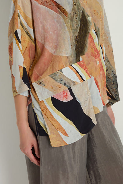 The person models the Abstract Earth Print Shirt, a loose-fit viscose-blend top with large buttons, paired with wide gray pants to create a relaxed silhouette against a plain light background.