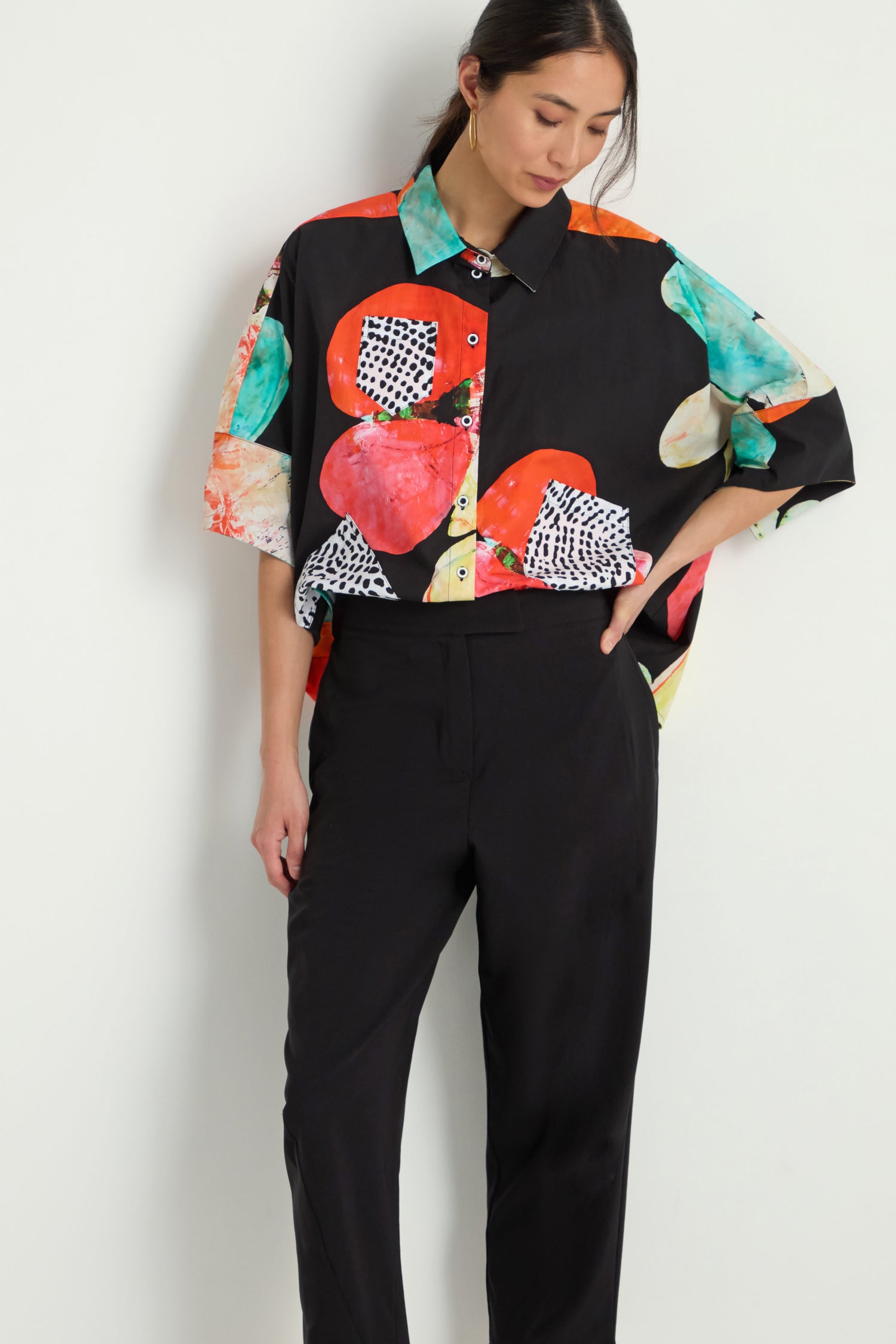 A person stands against a plain white background wearing Slim Leg Trousers in black and a loose, colorful button-up shirt with abstract patterns.