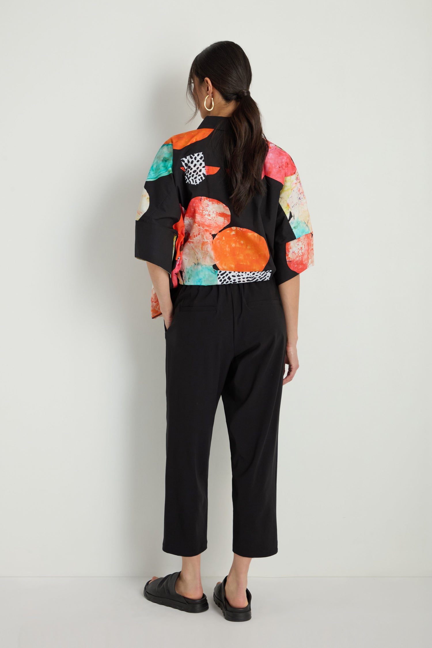 A person stands facing away, wearing a black top with colorful abstract patterns, Slim Leg Trousers in stretch fabric with an ankle-grazing length, black sandals, and large hoop earrings.