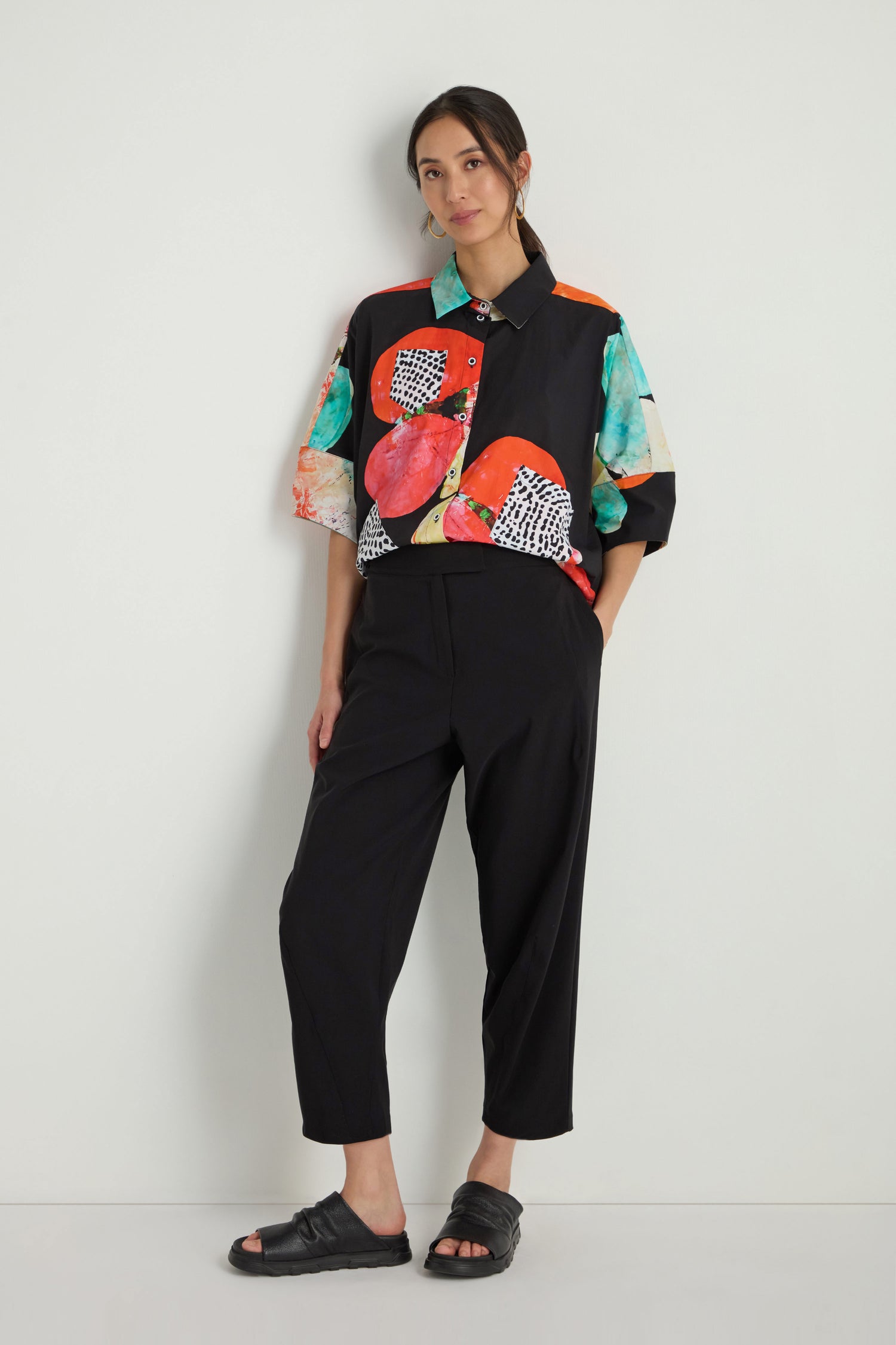 A woman stands against a white wall wearing a colorful patterned short-sleeve shirt, Slim Leg Trousers at ankle length, and black sandals.
