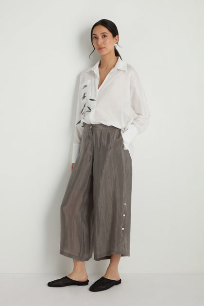 A woman stands against a white wall wearing a white embroidered shirt, black slip-on shoes, and the Wide Leg Button Trousers, made from a soft lyocell blend with side buttons for all-day comfort.