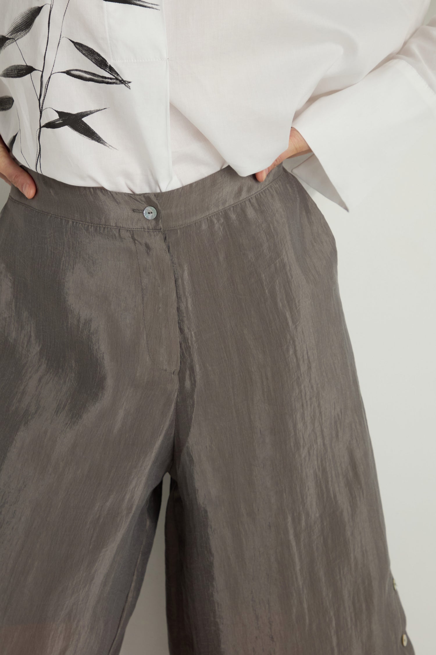 A person wearing high-waisted gray Wide Leg Button Trousers with a loose fit and a white shirt featuring a black leaf design on the left side.