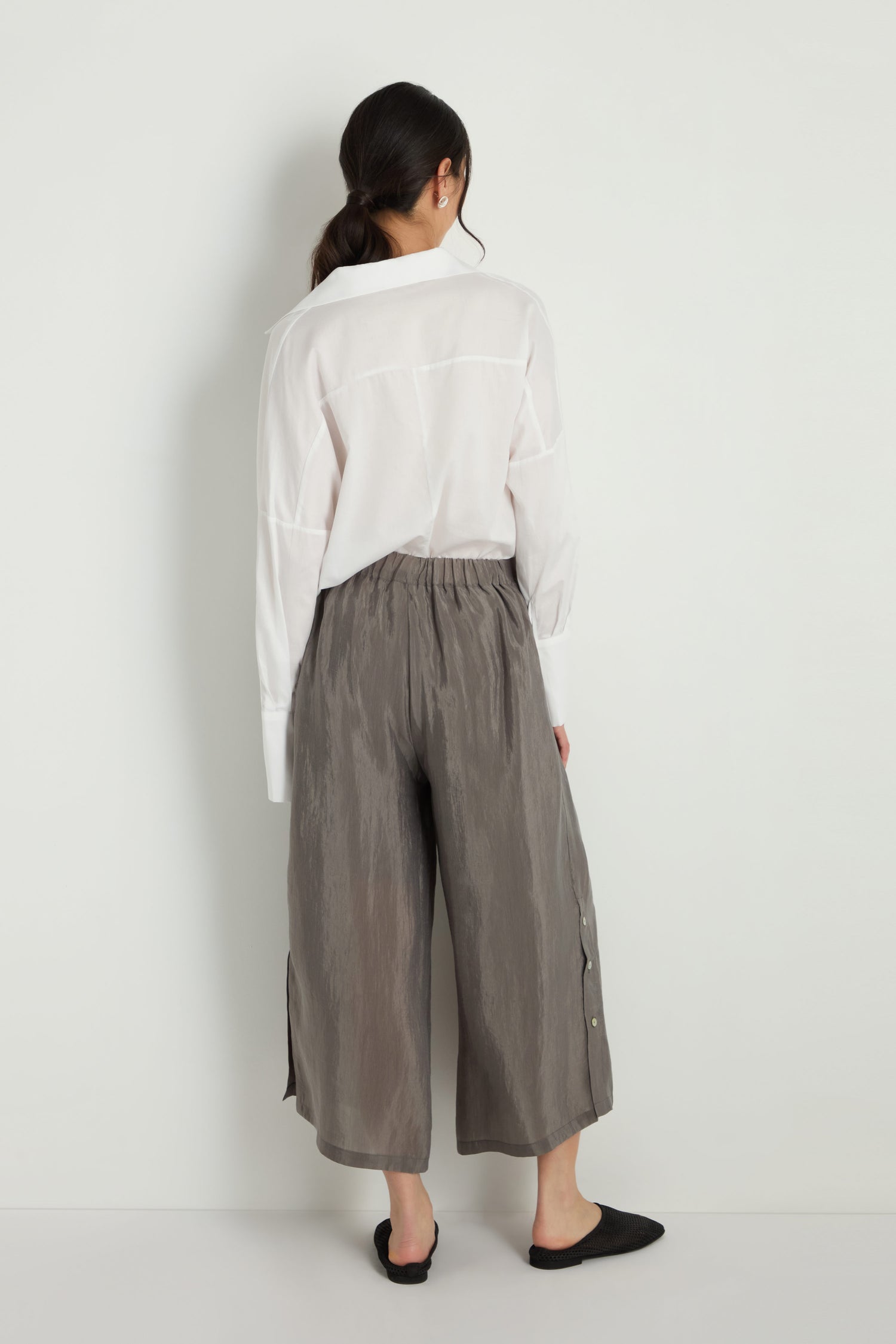 A woman stands against a white wall, facing away. She wears a white long-sleeve shirt, black flat shoes, and gray Wide Leg Button Trousers made from a lyocell blend.