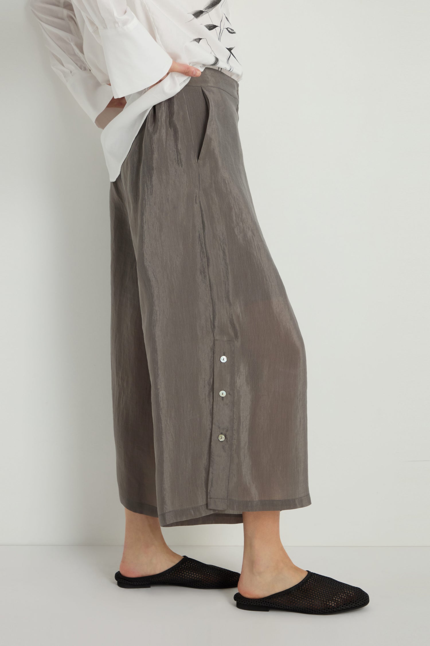 A person stands against a plain white background wearing Wide Leg Button Trousers in gray, crafted from a lyocell blend with side button details, paired with a white long-sleeve shirt and black mesh slip-on shoes.