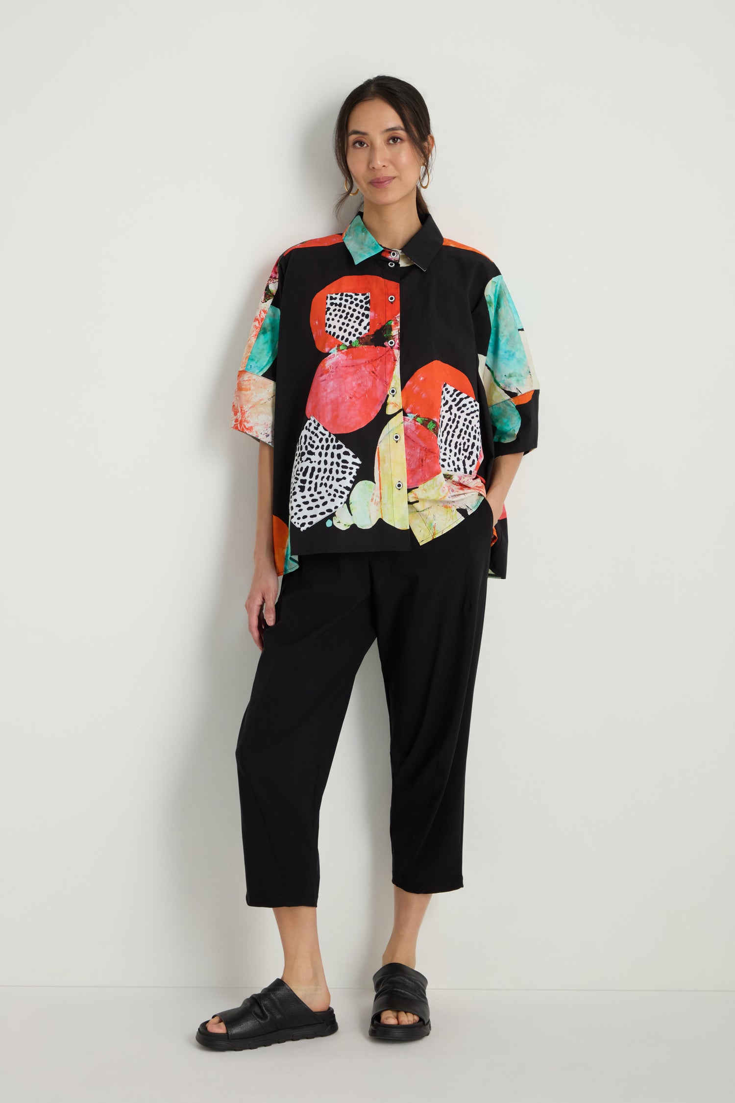 A woman stands against a white wall wearing the Bright Floral Boxy Shirt with a bold floral print, paired with black cropped pants and black sandals.