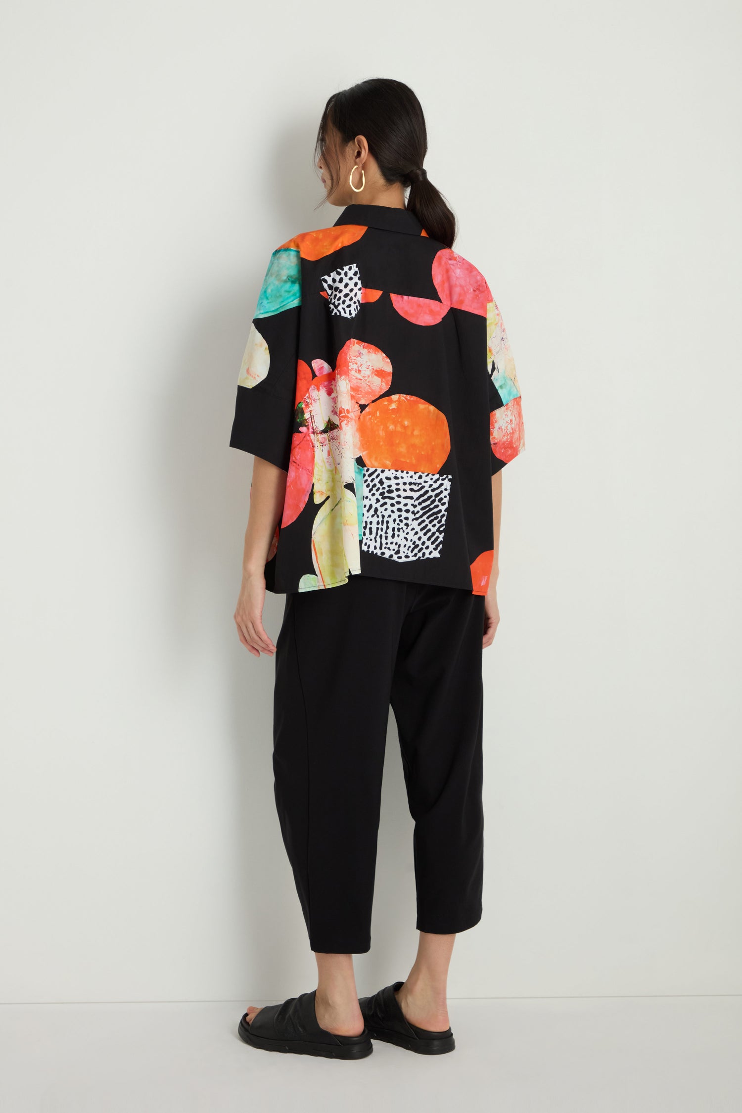 A woman stands facing away, wearing black cropped pants, black slide sandals, and the Bright Floral Boxy Shirt with colorful abstract patterns and white graphic details.