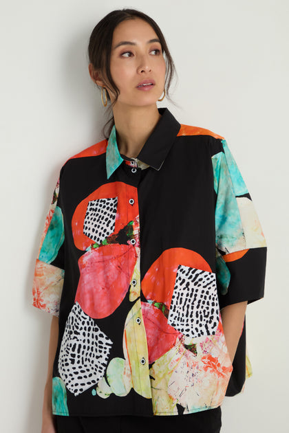 A woman wears the Bright Floral Boxy Shirt, featuring a loose, boxy fit and vibrant abstract floral pattern, as she stands against a plain light-colored wall.