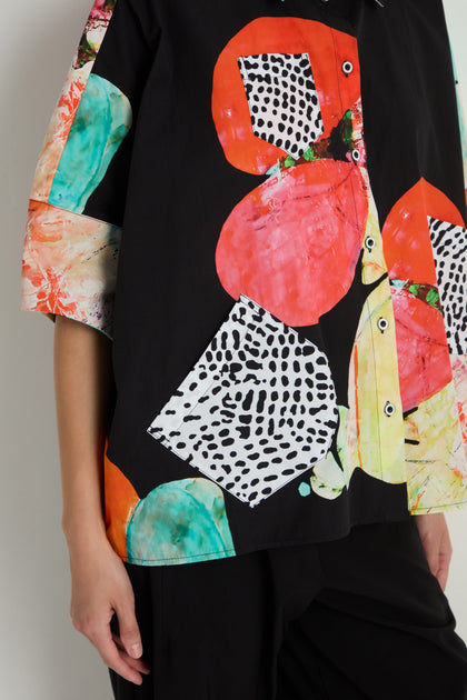 A person wearing the Bright Floral Boxy Shirt, a pure cotton black boxy shirt featuring colorful abstract shapes and patterns with red, yellow, and black-and-white geometric designs.