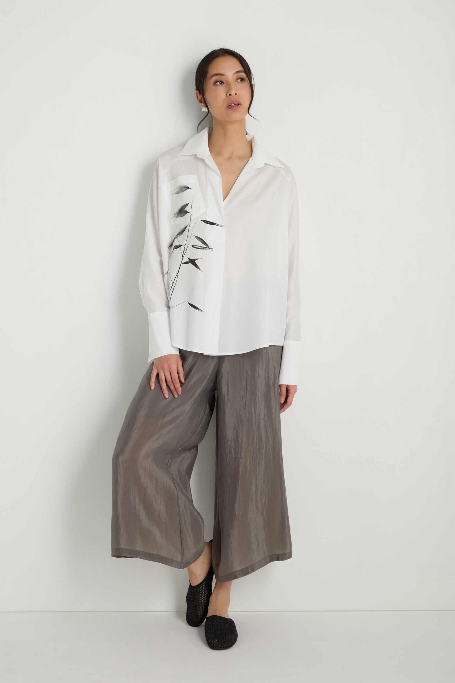 A woman stands against a white wall wearing a Bamboo Print Shirt, wide gray culottes, and black slip-on shoes.