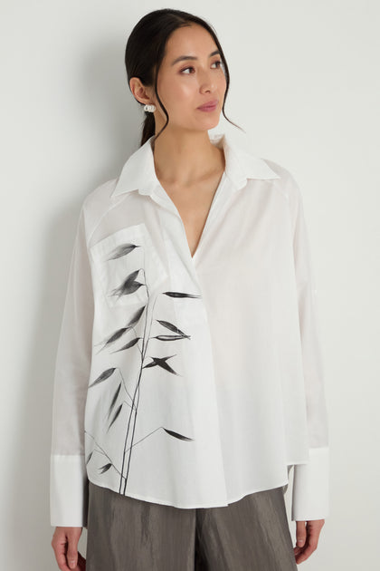 A woman stands against a white wall wearing the Bamboo Print Shirt with wide cuffs and loose gray pants, creating an effortlessly chic look.