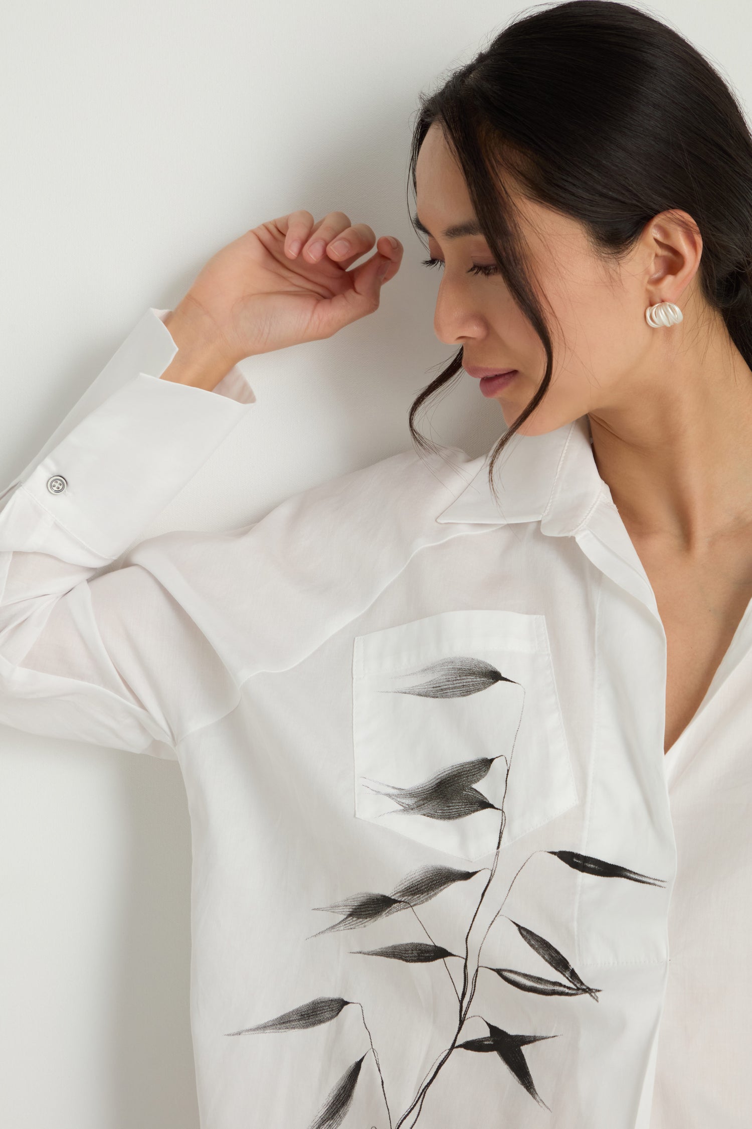 Woman in a pure cotton Bamboo Print Shirt with a black leaf design on the pocket, leaning against a white wall and looking down with her right arm raised.