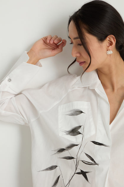 Woman in a pure cotton Bamboo Print Shirt with a black leaf design on the pocket, leaning against a white wall and looking down with her right arm raised.