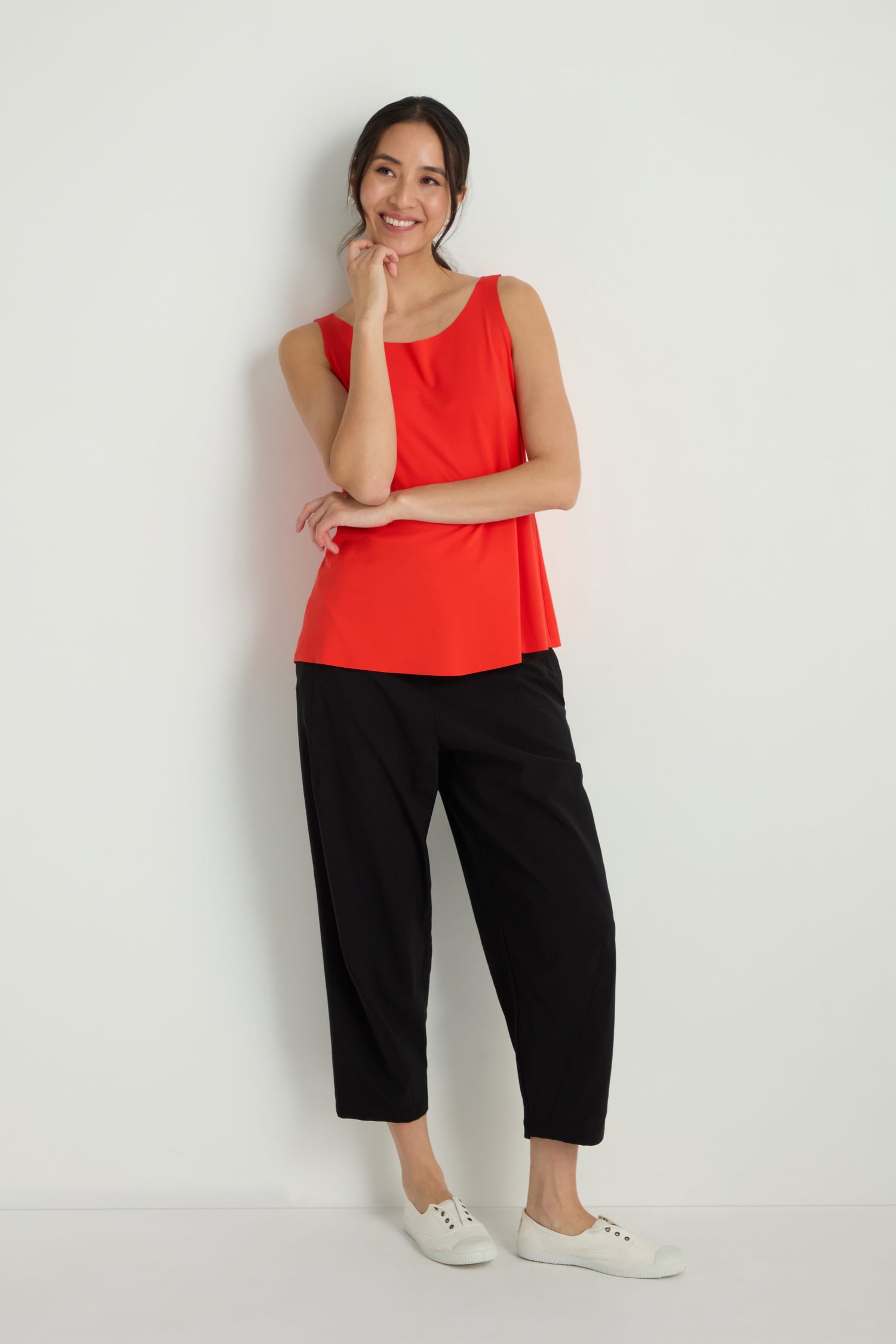 A woman stands against a white wall, wearing the Sleeveless Chilli Jersey Top with black pants and white sneakers. Her hair is tied back, and she gently touches her chin.