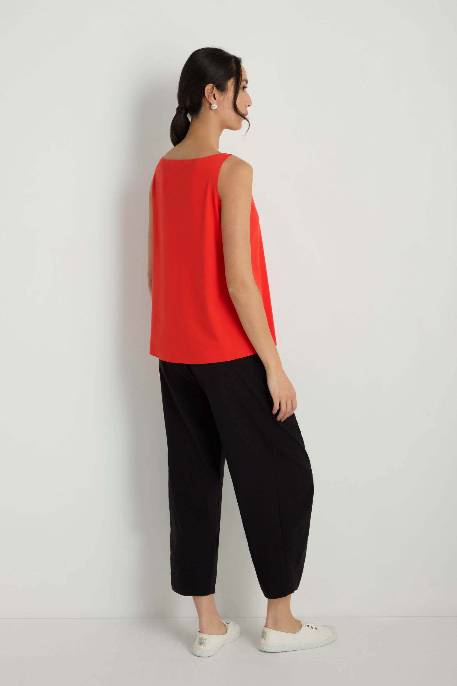 A woman stands sideways against a plain light wall, wearing the Sleeveless Chilli Jersey Top, loose black pants, and white slip-on shoes.